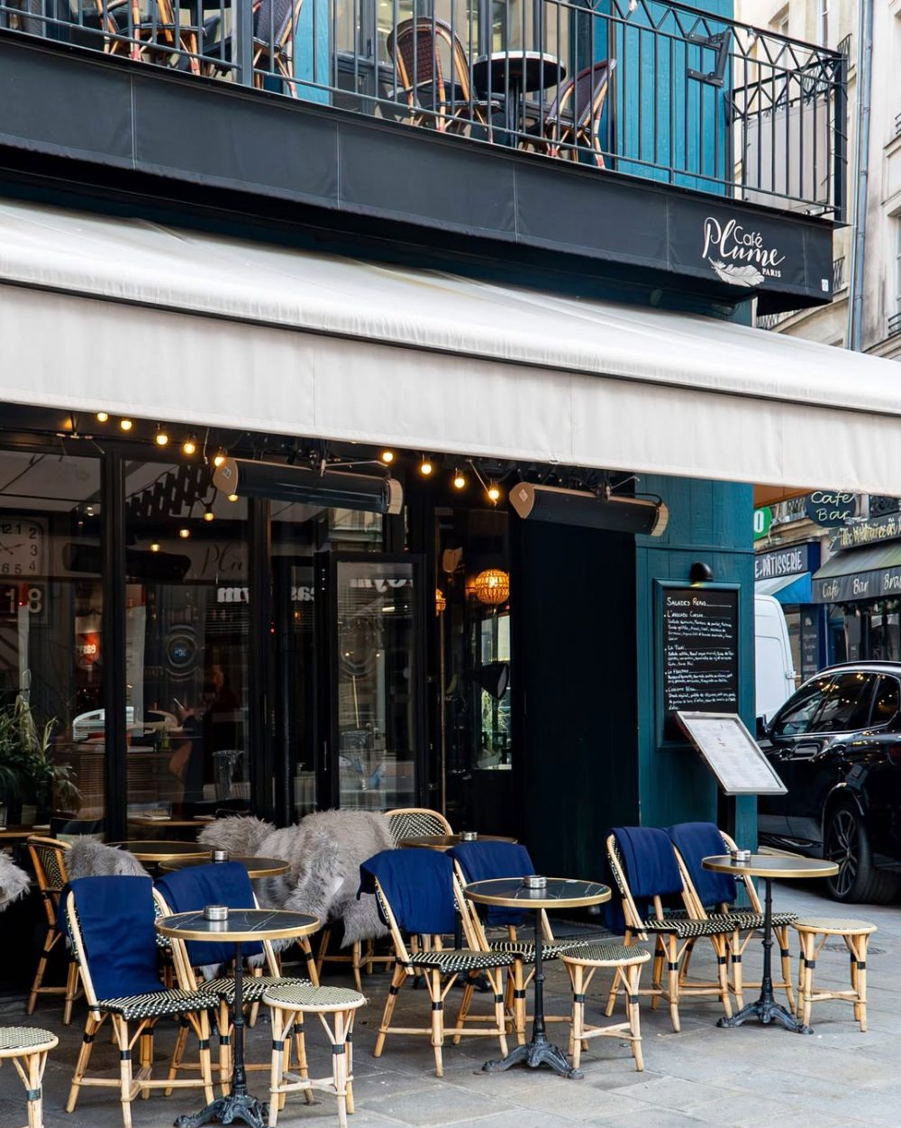 La façade du Café Plume : store blanc, guirlande d'ampoules, terrasse en rotin avec plaids bleus et balcon à l'étage