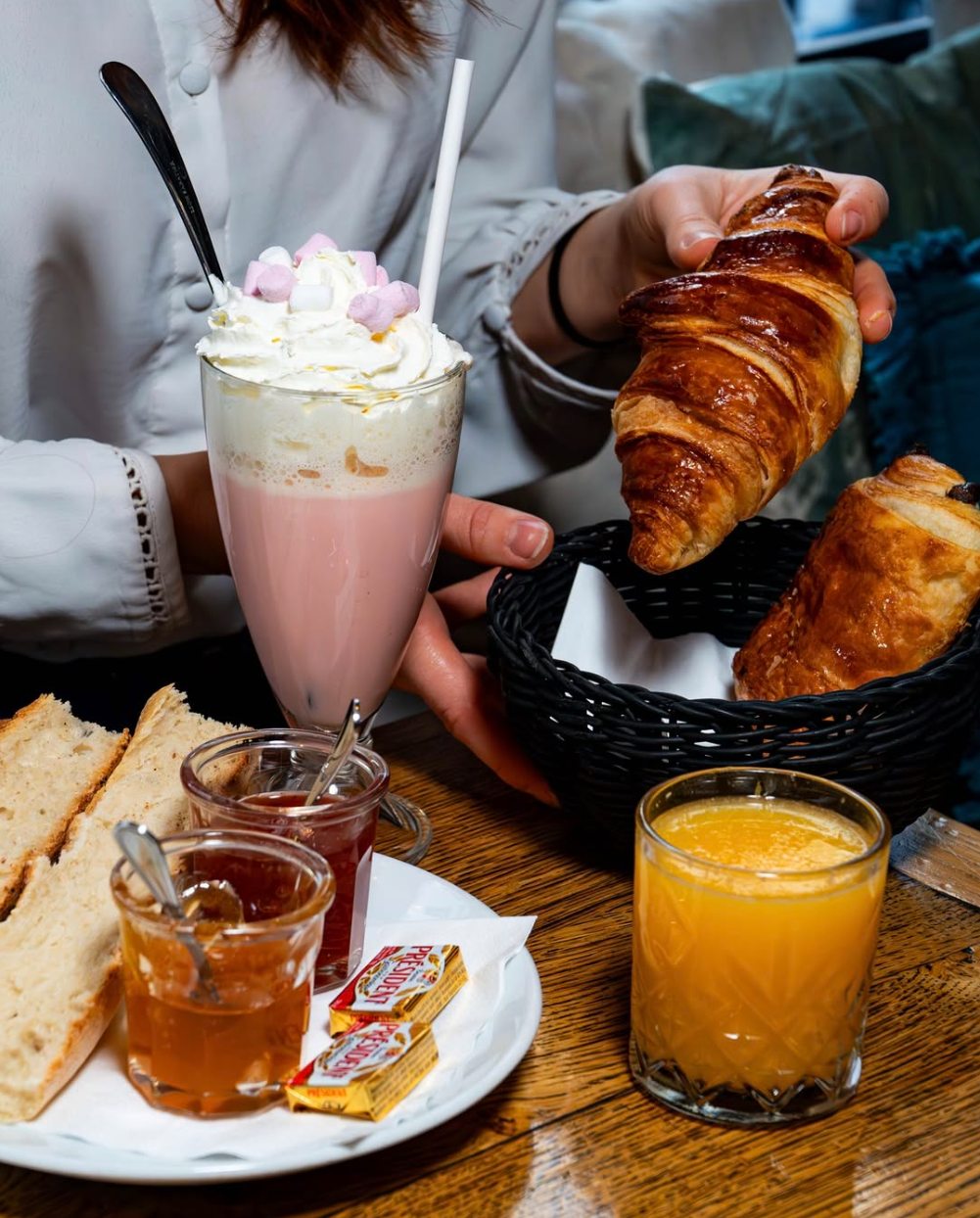 Petit-déjeuner sur une table en bois : croissant, pain au chocolat, tartines, confitures, jus d'orange et chocolat chaud à la chantilly